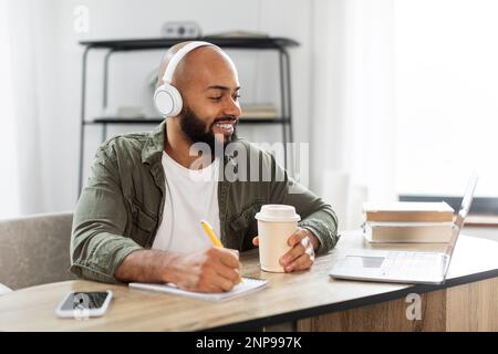 Happy latin man in headphones watching webinar remotely from home, drinking coffee and making notes in copybook Stock Photo