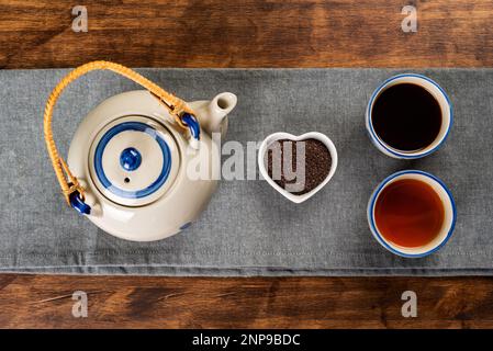 Porcelain teapot next to a small heart-shaped bowl with ground tea, two cups with different types of tea, darker red and lighter green. Stock Photo