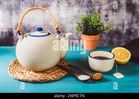Porcelain teapot next to a cup of tea and half a lemon, with two wooden spoons, one with white sugar and the other with brown sugar. Stock Photo
