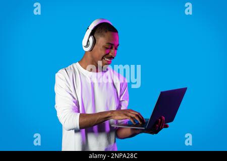 Joyful black guy using modern laptop and wireless headphones, gaming Stock Photo