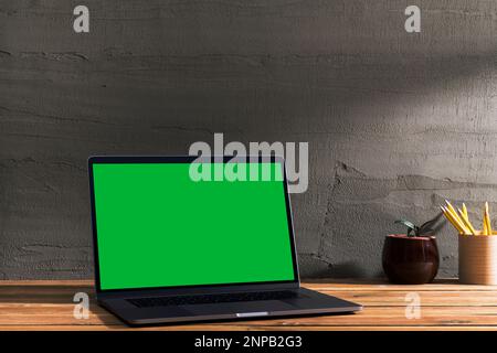 Chroma key green screen, angled view laptop on wooden table in front of concrete wall. Table top shot of interior space with window light effect. Stock Photo