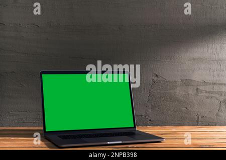 Chroma key green screen, angled view laptop on wooden table in front of concrete wall. Table top shot of interior space with window light effect. Stock Photo