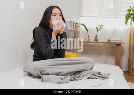 Asian female, young woman having cold, high temperature on bed and blowing her nose with tissue while suffering Stock Photo