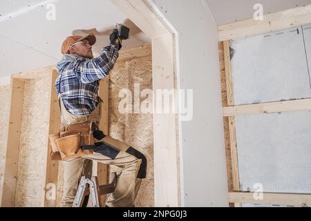 General Construction Contractor Attaching Drywall Using Cordless Drill Driver. Caucasian Remodeling Worker in His 40s. Stock Photo