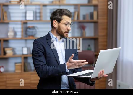 A mature male lawyer inside the office is standing at the desk, the businessman boss is talking remotely via video call using a laptop, man chatting with colleagues, advising online clients. Stock Photo