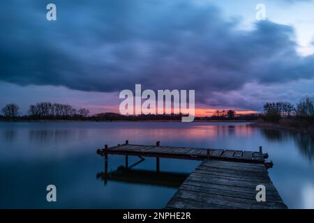 View from a wooden pier on a calm lake during sunset with dark rainy clouds Stock Photo