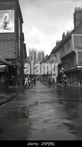 1950s, historical, distance view of York Minster from Low Petergate, York, England, UK. Founded in 627 AD, the Anglican cathedral is one of the world's most spectacular. A street in the city centre, Petergate is divided into High Petergate and Low Petergate. On the left, on a wall of a building is a billboard or poster of a young lady in a swmming costume and sun hat, with the line 'Ovaltine is Delicious & Refreshing'.  Also in the picture are signs for; Petergate Fish Restaurant, Merrimans, The Kettering & Leicester Boot Co & Surgical Stores. Stock Photo