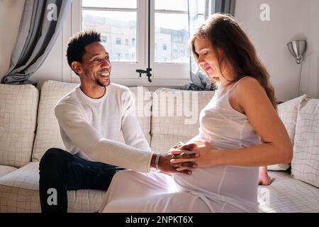 Man touching his pregnant wife's belly while sitting on the sofa at home. Stock Photo