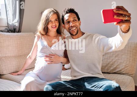Multiethnic couple expecting a baby taking selfies with a mobile phone at home. Stock Photo