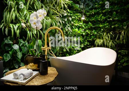 Bath in modern beauty salon, luxury bathroom interior in spa with vertical garden. Green plants wall, flowers and bathtub in hotel. Concept of nature, Stock Photo