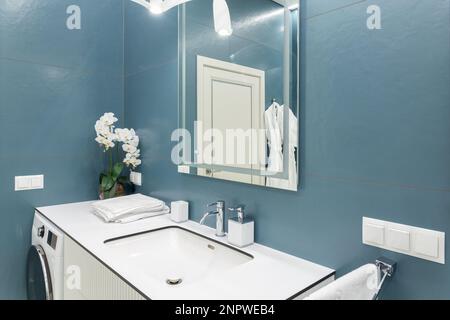 Blue and white bathroom with big mirror, towels, lighting, countertop basin and washing machine. Clean and fresh bathroom with modern light. Spacious Stock Photo