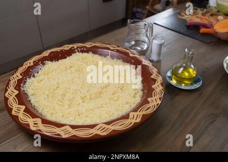Couscous grains in traditional bowl with water, salt, oil. Preparing food, ingredients for cooking process. Stock Photo