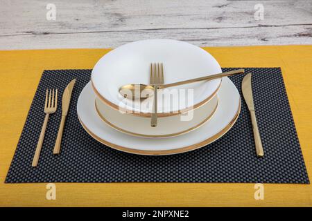 Plates and utensils, Plate, Bowl and golden cutlery against dining table background, side view. Modern craft ceramic tableware. Stock Photo