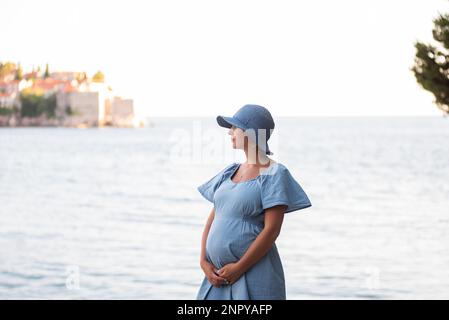 Pregnant woman traveler hugs baby in her belly near the island of Sveti Stefan in Montenegro. Close-up view middle aged millennial in blue denim dress Stock Photo