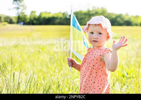 flag of Ukraine flutters in hands of little girl walking across field. Independence Day of Ukraine in village. Child waving Stock Photo