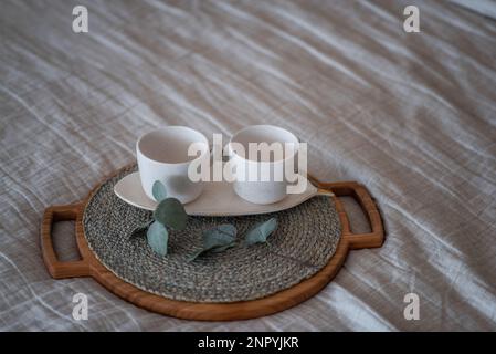On the Linen bedspread lies a wooden round tray, on it are two empty ceramic cups, green eucalyptus. Stylish, trendy morning. Textural, Scandinavian R Stock Photo