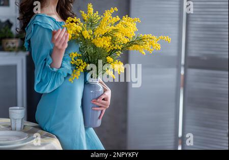 Faceless girl in a blue dress holds a vase with a bouquet of yellow mimosa in her hands. Spring, festive atmosphere. Allergy to flowers. Light loft in Stock Photo