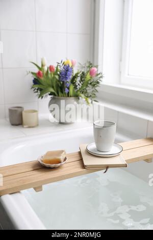 Cup of coffee, candles, book and soap on wooden board. Elegant Scandinavian bathroom interior with white tiles.Blurred background. Colorful tulips Stock Photo