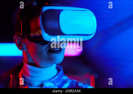 Neon metaverse futuristic concept. Portrait of trendy 40 years old woman in vr headset on dark background. Stock Photo