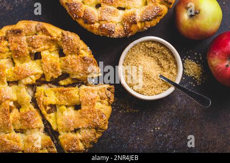 Top view of delicious homemade apple pie with cut slice, fresh apples, peel, cane sugar on brown rustic stone background. Autumn or Thanksgiving Stock Photo