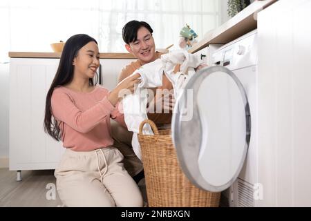 Young Asian married happy couple smiling and doing laundry at home. Boyfriend and girlfriend putting clothes in front loading washing machine together Stock Photo