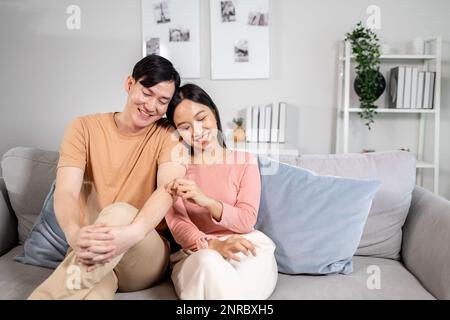 Young Asian married happy couple smiling and sitting on couch in living room at home. Boyfriend and girlfriend embracing and relaxing Stock Photo