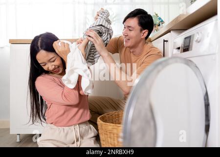 Young Asian married happy couple smiling and doing laundry at home. Boyfriend and girlfriend putting clothes in front loading washing machine together Stock Photo