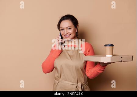 Pretty waitress talks on mobile phone, smiles at camera, poses with takeaway food in eco containers on beige background Stock Photo