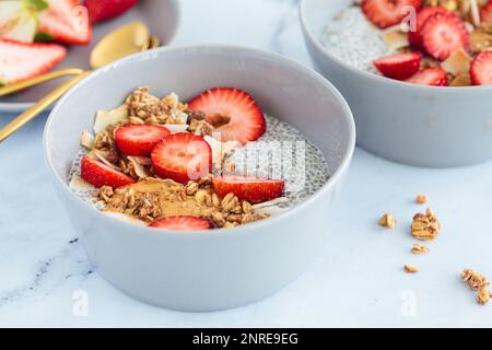 Chia pudding with homemade coconut granola, peanut butter and strawberries in gray bowl, marble background. Healthy plant based diet, detox, summer re Stock Photo