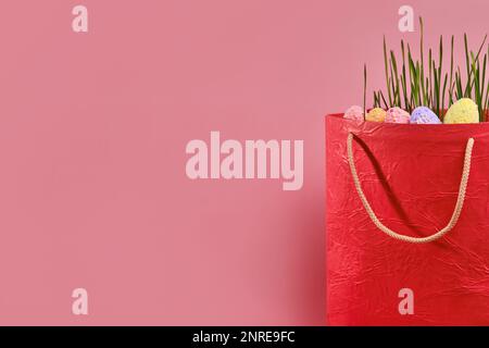 Red shopping bag, green grass and Easter eggs on pink background with copy space. Online shopping for the holidays. Symbols, traditions concept. Stock Photo