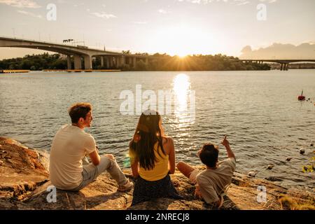 Man reclining and pointing at sea to friends sitting on rock by sea during sunset Stock Photo