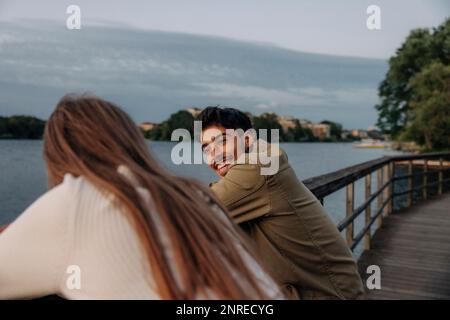 Side view of happy young man talking with female friend while leaning on railing at pier Stock Photo