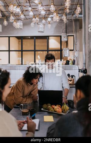 Male and female programmers discussing over laptop by colleagues in cafeteria at office Stock Photo