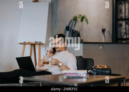 Businessman having coffee while talking on smart phone and working over laptop in office Stock Photo