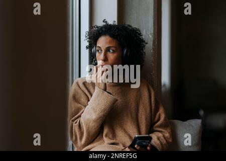 Contemplative businesswoman with smart phone listening to music through wireless headphones in office Stock Photo