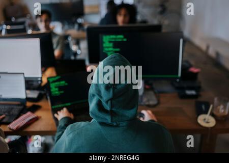 Rear view of young male hacker wearing hood coding on computer at desk in creative workplace Stock Photo