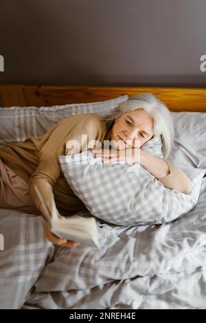 Senior woman reading book while lying on bed at home Stock Photo