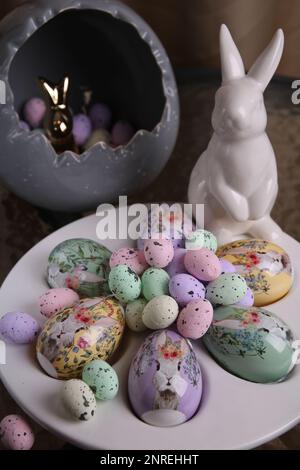 photo a lot of bright decorative large and small eggs lying on a white plate and next to it is a white rabbit on a glass table Stock Photo