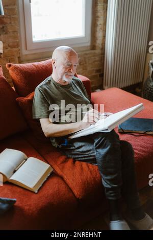 Senior man doing painting while sitting on sofa by book at home Stock Photo