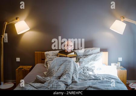 Senior man spending leisure time while reading book on bed at home Stock Photo