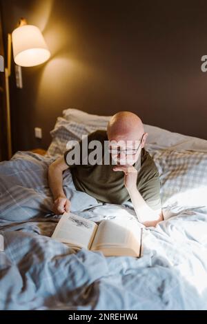 Senior man with hand on chin reading book while lying on bed at home Stock Photo
