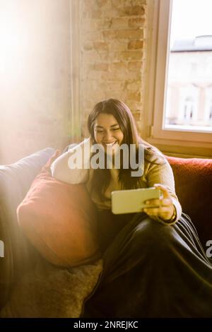 Happy woman taking selfie through mobile phone sitting on sofa at home Stock Photo