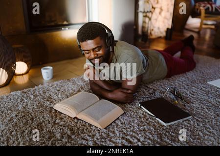 Smiling young man listening to music and reading book while lying on carpet at home Stock Photo
