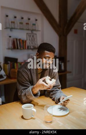 Smiling man using smart phone and having doughnut at home Stock Photo