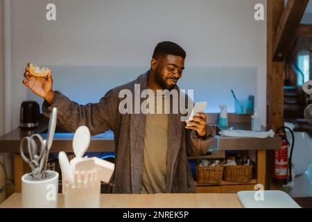 Happy man holding doughnut and using internet on smart phone at home Stock Photo