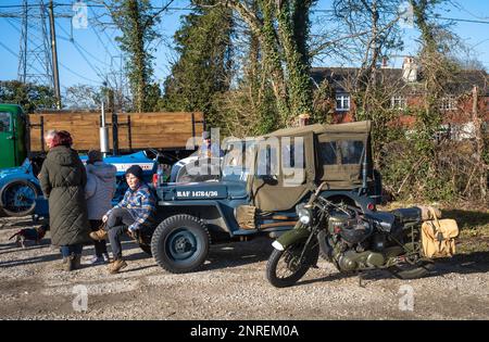 A boy sits on the front bumper of a WW2-era Jeep painted in RAF livery that is parked next to a WW2-era Norton motorcycle.at a vintage vehicles event Stock Photo