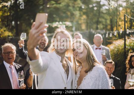 Happy groom taking selfie with bride and guests through smart phone at wedding ceremony Stock Photo