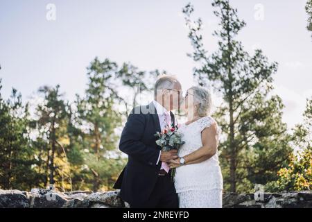 Affectionate newlywed senior couple kissing on mouth against forest on wedding day Stock Photo