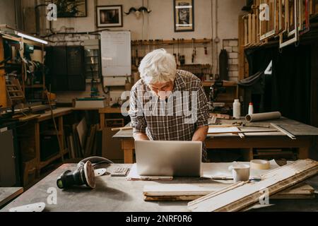 Senior male carpenter holding pencil in mouth while working on laptop at workshop Stock Photo