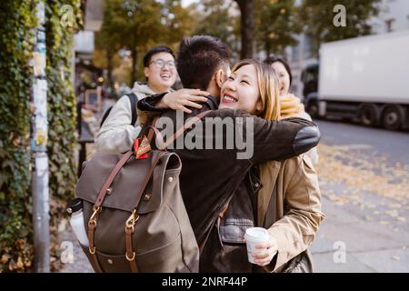 Happy young woman embracing male friend wearing backpack while standing at sidewalk Stock Photo